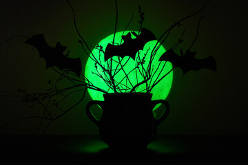 Mysterious black cauldron with dry plants and cutout bats in front of a green moon background....