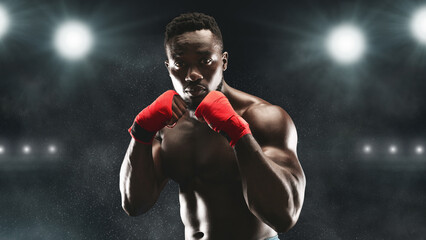 A boxer in red hand wraps stands ready in a dark stadium, illuminated by bright lights.