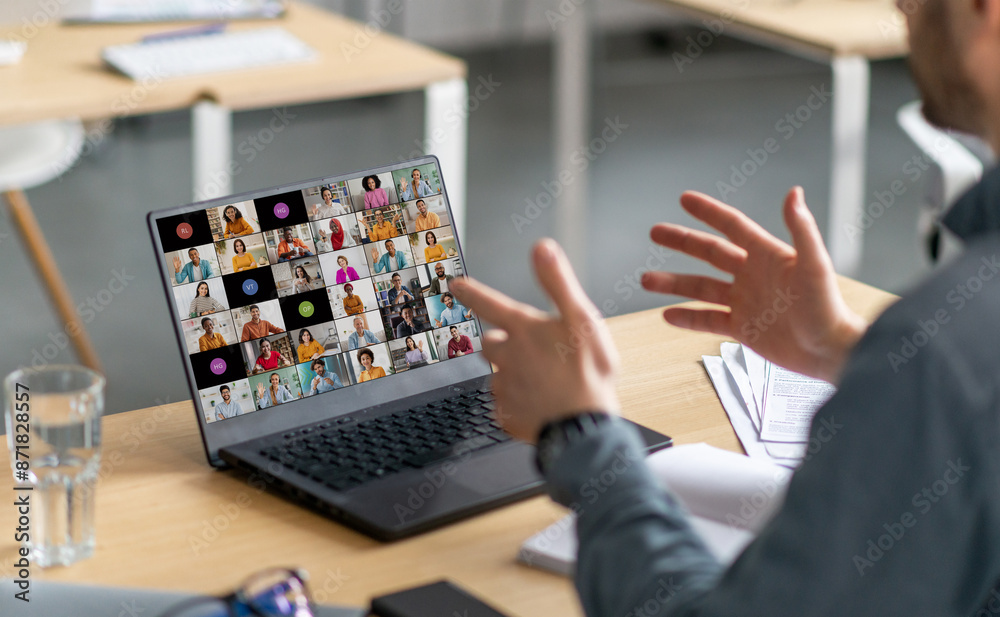 Poster A man sits at a desk in front of a laptop. He is gesturing with his hands and looking at the screen, which displays a grid of video calls with multiple people.