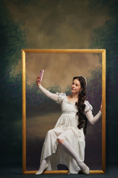 Social media event promotion. Beautiful young one in Victorian white dress sitting in picture frame and taking selfie with mobile phone on vintage background. Concept of history, comparison of eras