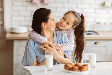 A young girl with long brown hair smiles and hugs her mother while they sit together at a kitchen table. They are both looking at each other.