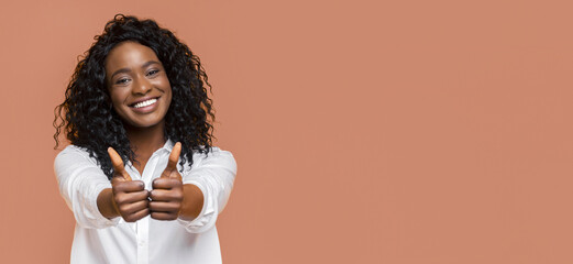 A young black woman with curly black hair smiles brightly at the camera, giving two thumbs up with...
