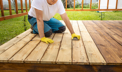 A man in yellow gloves sands a wooden deck in preparation for staining or painting. The deck is...