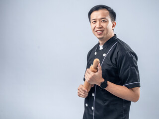 Fototapeta premium A smiling Asian male chef in a black chef's uniform, holding a wooden rolling pin, exudes friendliness and readiness for baking tasks against a light blue background
