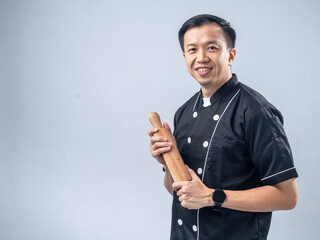 A smiling Asian male chef in a black chef's uniform, holding a wooden rolling pin, exudes friendliness and readiness for baking tasks against a light blue background