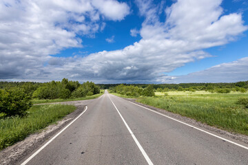 A straight, paved road winds through a lush green field, under a partly cloudy blue sky..