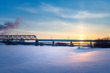Obraz premium A large bridge spans a frozen river at sunset, with the setting sun casting a warm glow over the snowy landscape.
