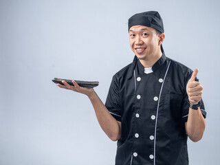 A smiling Asian male chef in a black chef's uniform and bandana, holding a stone sharpening tool in one hand and giving a thumbs-up, exudes confidence and positivity against a light blue background