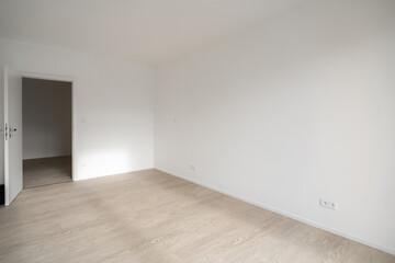 
A minimalist, empty room with white walls, light wooden flooring, an open door, and ample natural light.