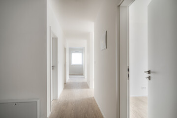 Minimalist, well-lit hallway in a modern apartment with wooden floors, white walls, and several doors leading to different rooms.