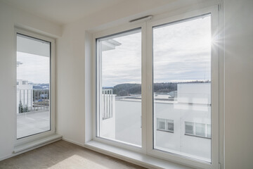 bright room with large floor-to-ceiling windows that offer a view of modern buildings and a distant forested area. The room has wooden flooring and white walls.