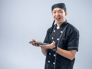 A smiling Asian male chef in a black chef's uniform and bandana, holding a stone sharpening tool with both hands, showcases his readiness and skill in the kitchen against a light blue background