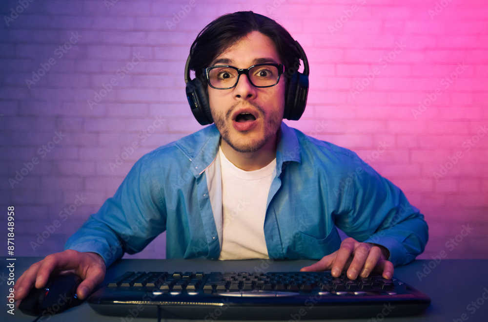 Poster a young man wearing headphones and glasses sits in front of a keyboard and mouse, looking surprised 