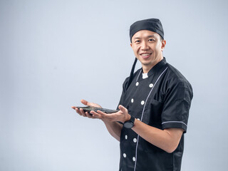 A smiling Asian male chef in a black chef's uniform and bandana, holding a stone sharpening tool with both hands, showcases his readiness and skill in the kitchen against a light blue background