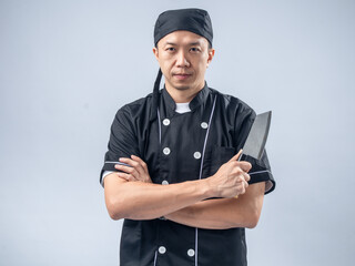 A confident Asian male chef in a black chef's uniform and bandana, holding a cleaver with arms...