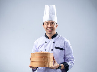 A smiling Asian male chef in a white chef's uniform and hat, holding a bamboo steamer and its lid,...
