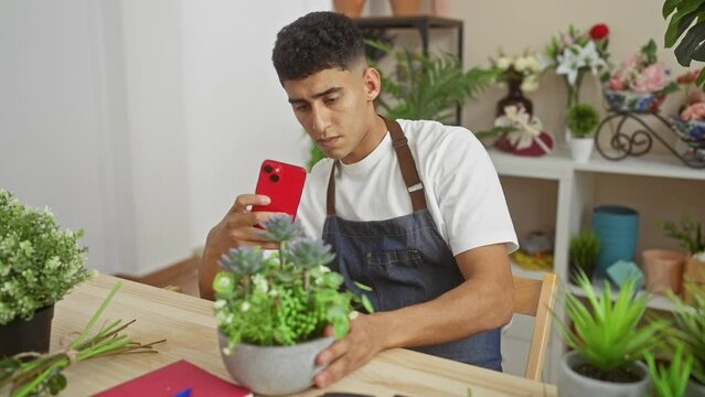 A young man captures a plant with his smartphone in an indoor flower shop filled with various greenery.