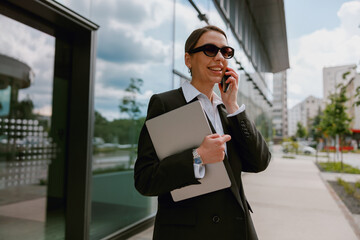 A businesswoman is standing outside a modern office, holding a laptop and talking on the phone