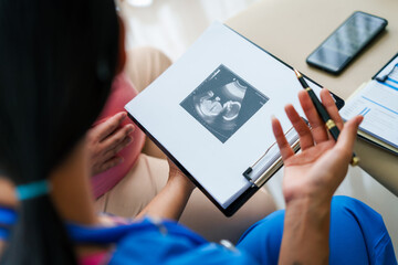 Asian female doctor and nurse consult with a pregnant woman on a sofa, using an ultrasound and stethoscope, preparing her for birth ahead of schedule with care and expertise.