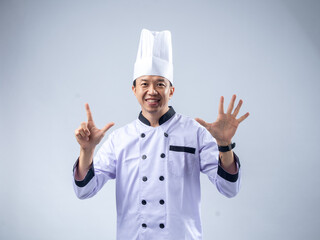 A smiling Asian male chef in a white uniform and hat holds up seven fingers, signifying the number seven, against a light blue background. The setting is professional and engaging