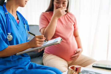 Asian female doctor and nurse consult on sofa with a pregnant woman, using ultrasound and...
