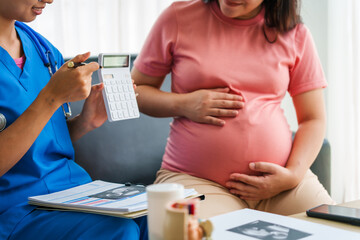 Asian female doctor and nurse consult on a sofa with a pregnant woman, using a calculator to discuss medical expenses, and using ultrasound and stethoscope for thorough examination.