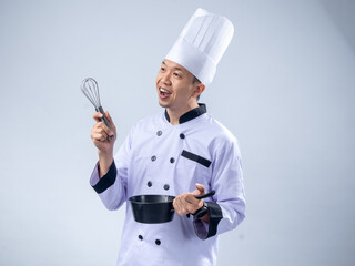 A cheerful Asian male chef in a white chef's uniform and hat, holding a whisk in one hand and a...
