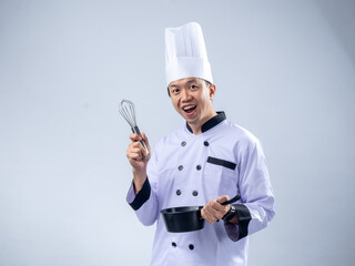 A cheerful Asian male chef in a white chef's uniform and hat, holding a whisk in one hand and a...
