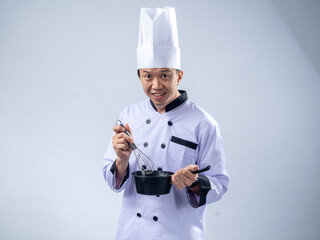 A cheerful Asian male chef in a white chef's uniform and hat, holding a whisk in one hand and a small pan in the other, smiles excitedly against a light blue background