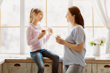 A mother and her young daughter are enjoying a snack together by a window in their home. The mother is standing and holding a glass of milk, while the daughter is sitting on a windowsill