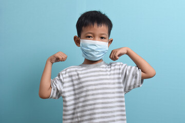 Asian Boy wearing health mask isolated on blue background. Confident kid shows his biceps arm muscle. Medical and health concept