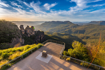 A panoramic view of the Three Sisters rock formation in the Blue Mountains, Australia. The photo was taken from the scenic Echo Point Lookout with a railing overlooking the vast landscape.