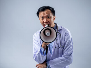 Asian male doctor in a white coat holding a megaphone and speaking enthusiastically. The plain background emphasizes the focus on the doctor's energetic announcement and the stethoscope