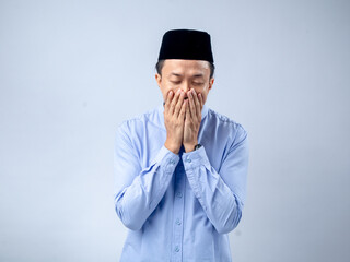 Asian man in traditional black cap and light blue shirt, with hands covering his face in a gesture...