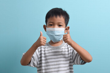 Asian Boy wearing a health mask while giving a thumbs up isolated on a blue background. Medical and health concept. Human population viruses, infections, flu disease