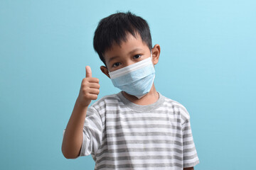 Asian Boy wearing a health mask while giving a thumbs up isolated on a blue background. Medical and health concept. Human population viruses, infections, flu disease