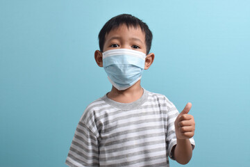 Asian Boy wearing a health mask while giving a thumbs up isolated on a blue background. Medical and health concept. Human population viruses, infections, flu disease