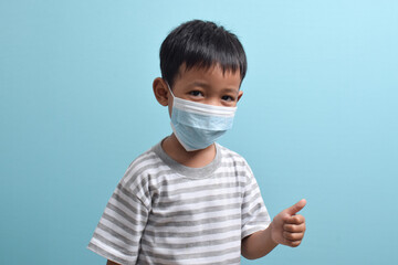 Asian Boy wearing a health mask while giving a thumbs up isolated on a blue background. Medical and health concept. Human population viruses, infections, flu disease