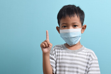 Asian Boy wearing a health mask while pointing his hand up isolated on blue background. Medical and health concept. Human population viruses, infections, flu disease