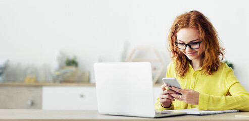 Happy businesswoman using smartphone near computer in office, surfing internet on phone