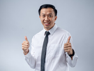A cheerful Asian male in a white shirt and tie, giving two thumbs up, expresses positivity and approval against a light blue background. The setting is professional and upbeat.