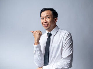 An Asian man in a white shirt and tie points to his side with a confident smile, standing against a light grey studio background. His professional attire and engaging gesture