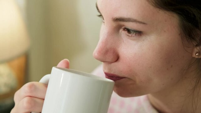Close-up portrait of an attractive young adult brunette woman in pajamas taking a sip of coffee from a mug in the kitchen on a sunny morning and looking straight ahead out the window