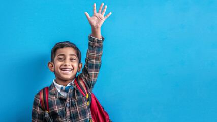 A smiling schoolboy in a checkered shirt and red backpack is energetically raising his hand against a blue wall in a classroom, showing enthusiasm for learning