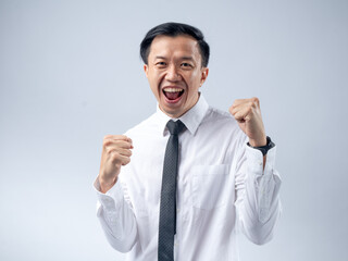 Asian businessman in white shirt and black tie celebrating success with fists clenched and mouth open in excitement, standing against a plain light background. His joyful expression and victory pose