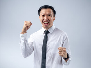 Asian businessman in white shirt and black tie celebrating success with fists clenched and mouth open in excitement, standing against a plain light background. His joyful expression and victory pose