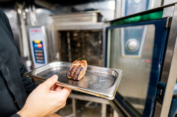 Close-up of a hand holding a metal tray with a single cooked steak in front of an industrial kitchen oven