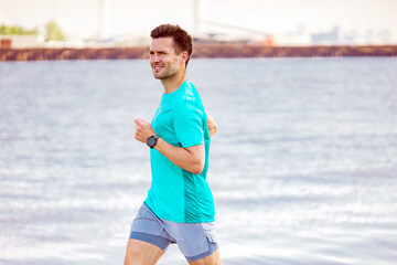 Joyful man jogging along the waterfront in a turquoise shirt, with a cityscape in the distant background.
