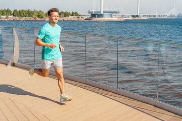 Man in turquoise shirt jogging on wooden deck by the sea with cityscape in the distance.