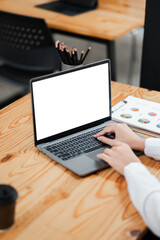 Fototapeta premium Hands typing on a laptop with a blank screen in a modern office setting, featuring a wooden desk, clipboard with graphs, and a coffee cup.